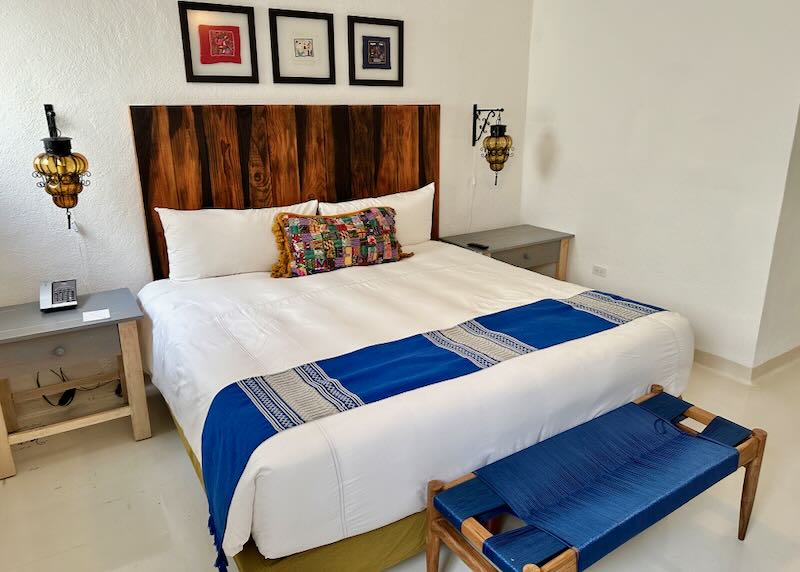 A white bed has a cobalt blue throw and matching woven bench at its foot.