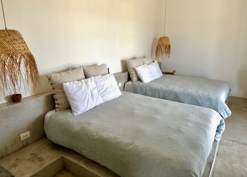 Two beds on a raised concrete platform sit in a hotel room with two woven basket lamps on each side.