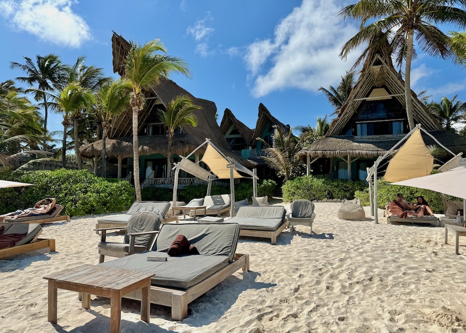 Double-sized sunbeds on the beach in front of a hotel made of a group of A-frame buildings with thatched roofs at Ahau hotel in Tulum.