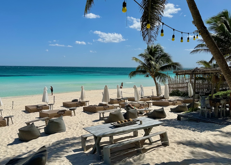 A wide, sandy beach with sunbeds, picnic tables, and palm trees at Casa Violeta in Tulum.