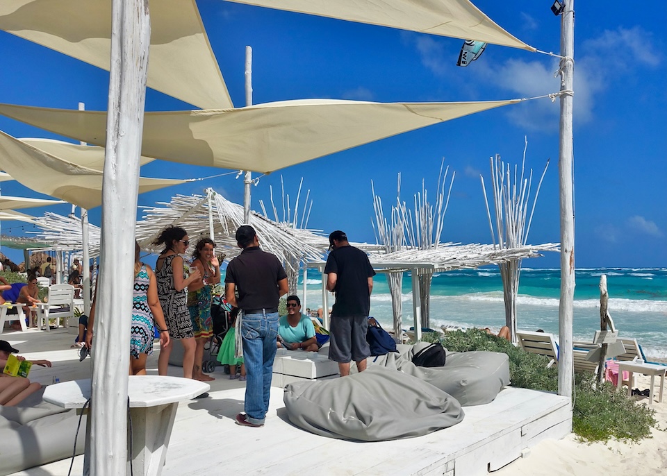 A group of people talking on a white, wooden deck with white sails above and a white bamboo pergola in front facing the ocean at Coco hotel in Tulum.