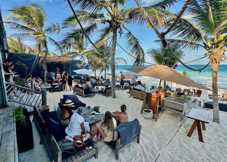 A busy beach club with people dining and drinking under palm trees with a disco ball at Casa Gitano in Tulum.