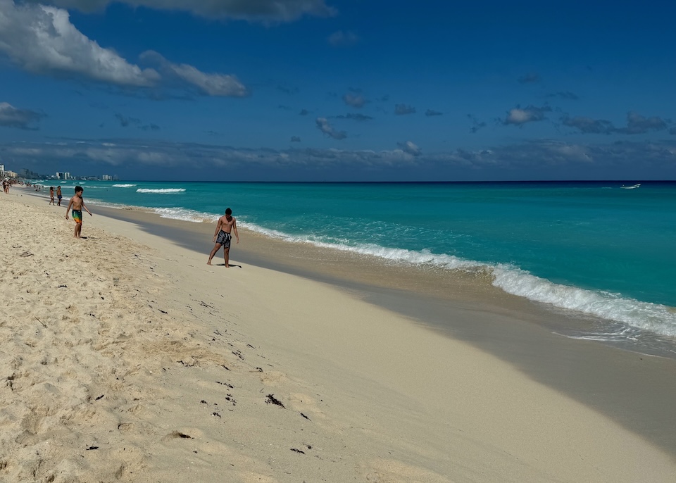 A wide stretch of sand on the beach with a clear ocean and a few people walking in the South Hotel Zone of Cancun