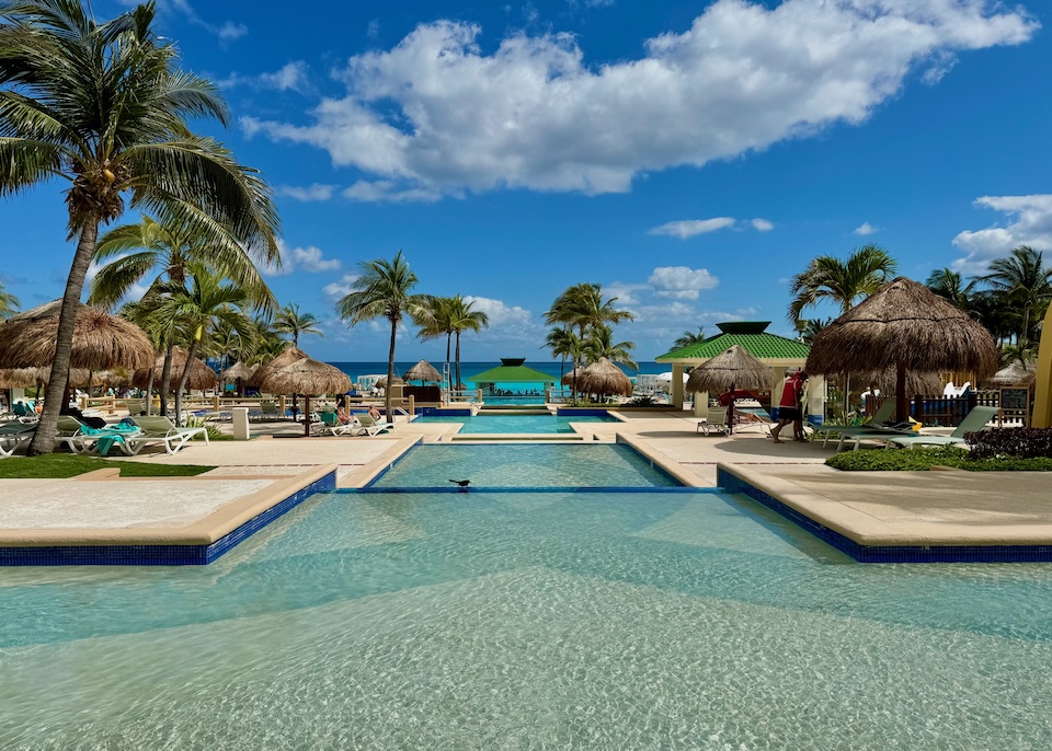 A series of shallow pools with palm trees and thatched umbrellas facing the ocean at Iberostar Selection in Cancun.