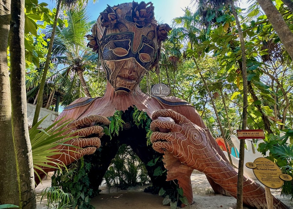 Facing the colossal wooden statue, Ven a la Lux, a bust of a woman with a flower crown and her hands holding open her chest, which is filled with ferns and vines in the Middle Beach Zone of Tulum.