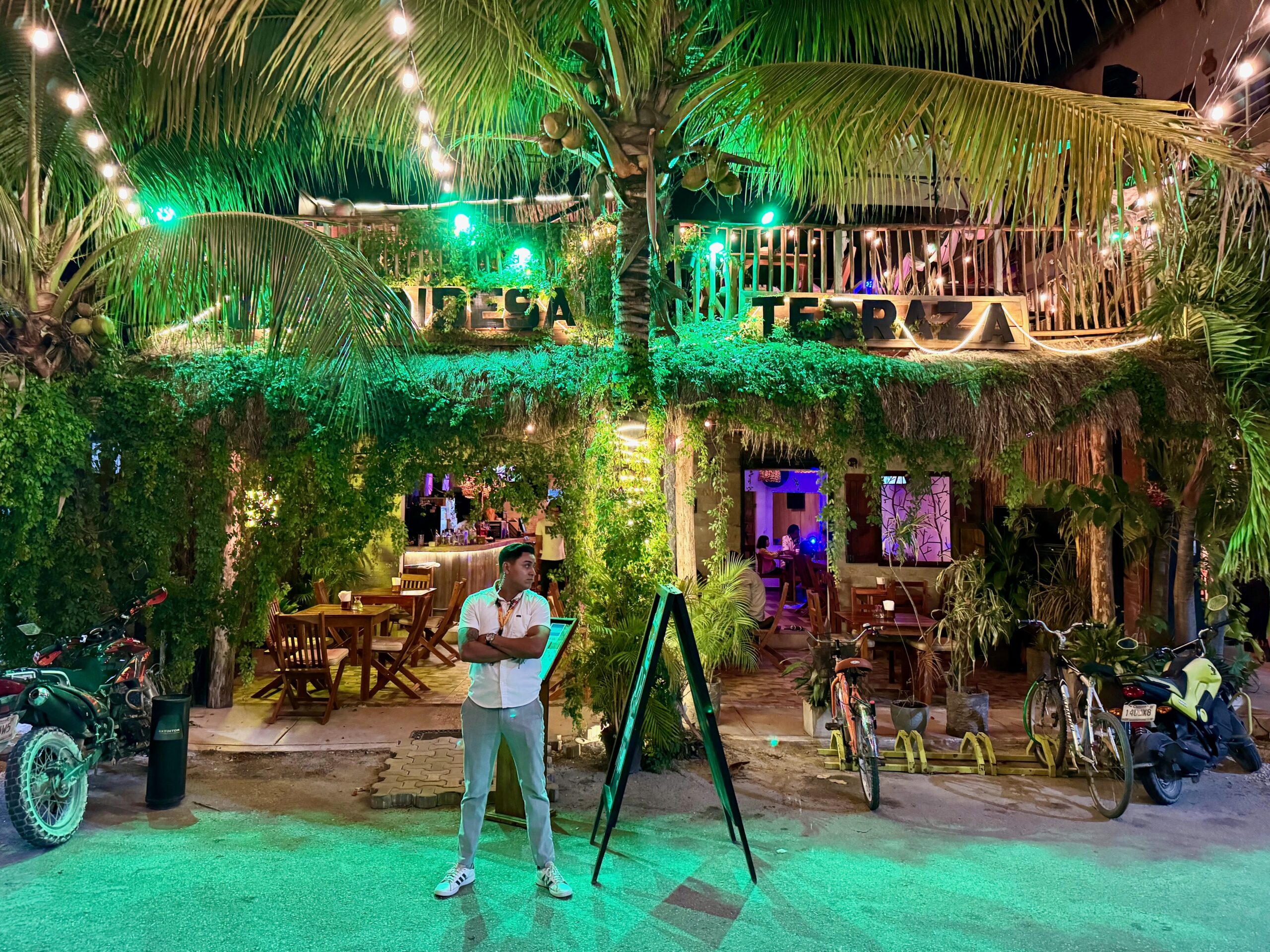 Outside a two-floor bar and restaurant covered in vines and foliage with palm trees lit up with fairy lights at night in Tulum pueblo.