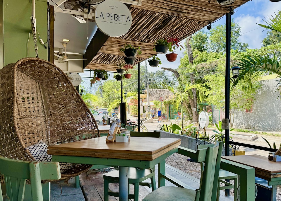 A sidwalk cafe with a latilla ceiling, hanging plants, an rattan egg-shaped chair, and a few tables in the La Veleta neighborhood of Tulum.