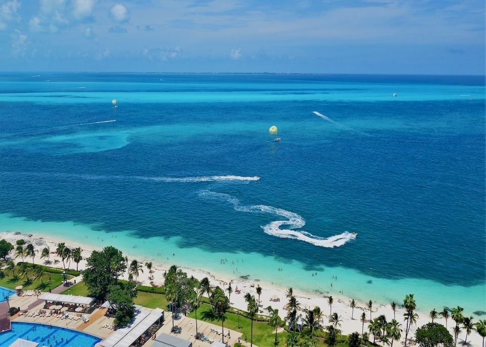 Jet skis are followed by their swirling, white, sea foam wakes, plus two parasailers glide above the ocean in front of the beach in the North Hotel Zone of Cancun.