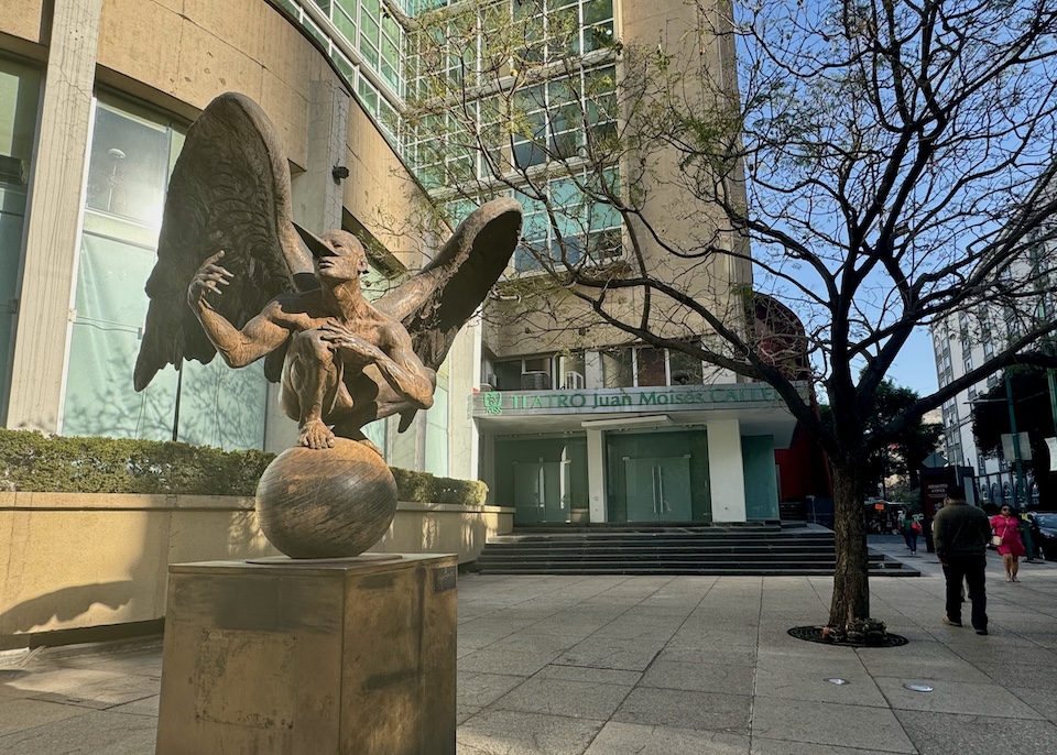 A statue of a man with wings and a pointy beak crouching on a globe in front of a theater in the Juarez neighborhood of Mexico City.