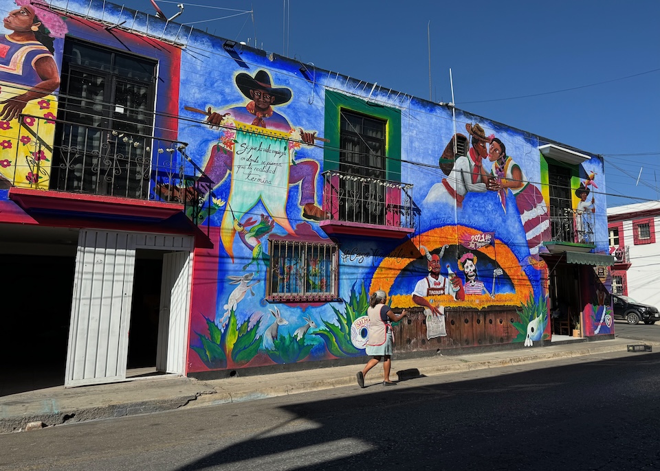 A woman in a cobbler apron walks on the sidewalk past a vibrant mural in Barrio de la Noria in Oaxaca