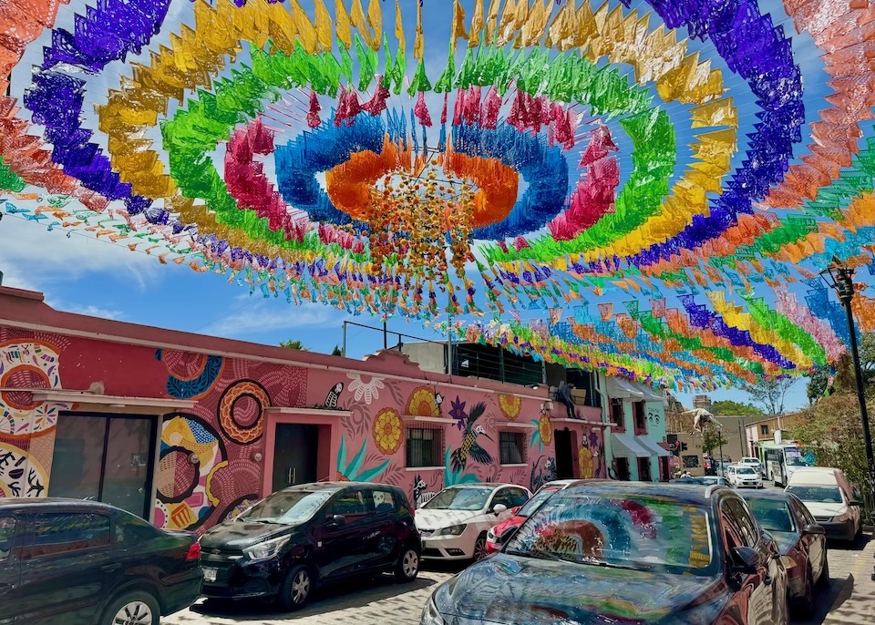 Colorful papel picado streamers hanging in over a street with floral murals on the sides of buildings in the Jalatlaco neighborhood of Oaxaca.