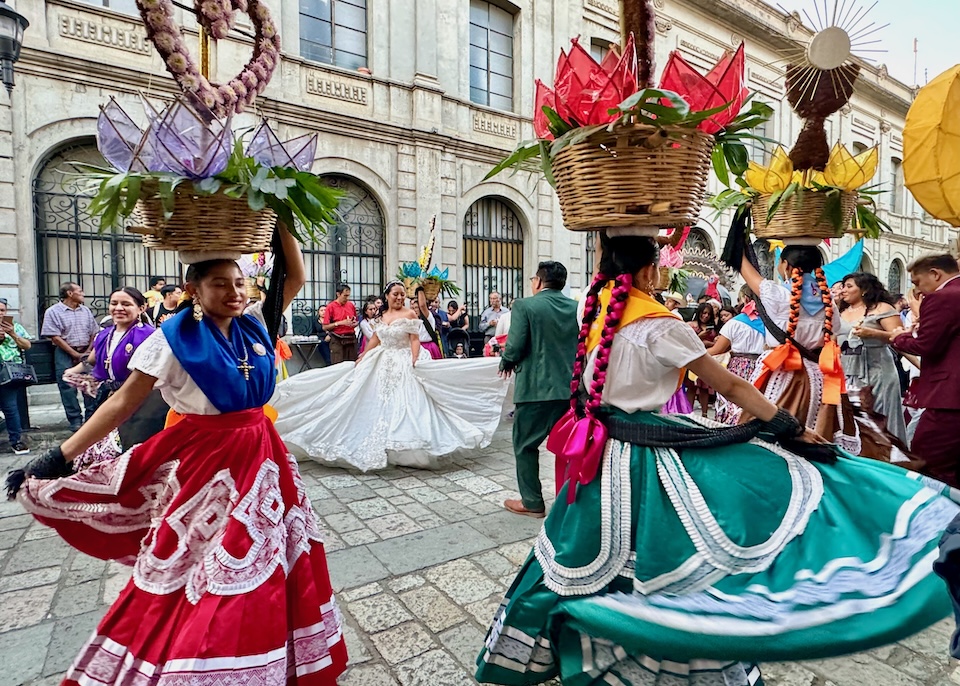 A bride in white and women in vibrant, traditional skirts hold baskets on their heads while dancing in the Santo Domingo neighborhood of Oaxaca.