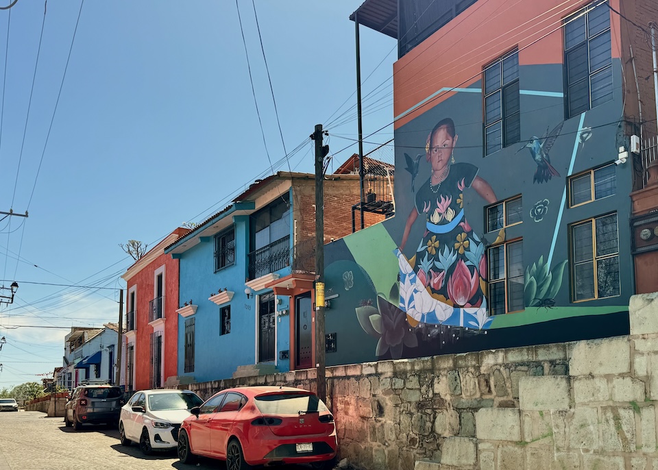 A mural showing a young girl in a floral dress with birds in a row of colorful buildings on a split-level street in the Xochimilco neighborhood of Oaxaca.