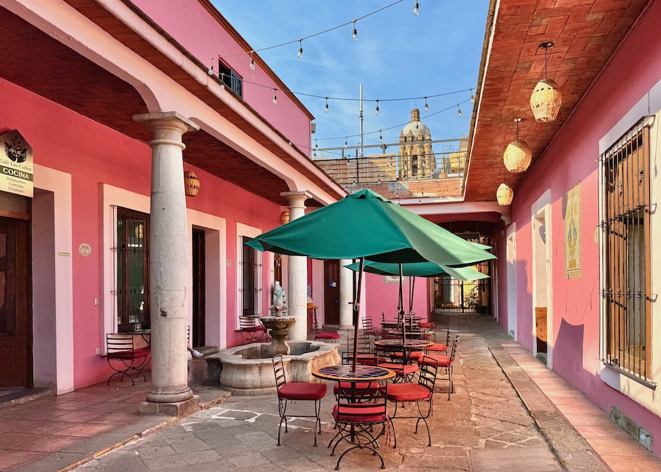 A pink interior courtyard with lights strung overhead and a bell tower of the Baroque church of Santo Domingo de Guzmán visible in the background at Hotel Pan:AM in Oaxaca.