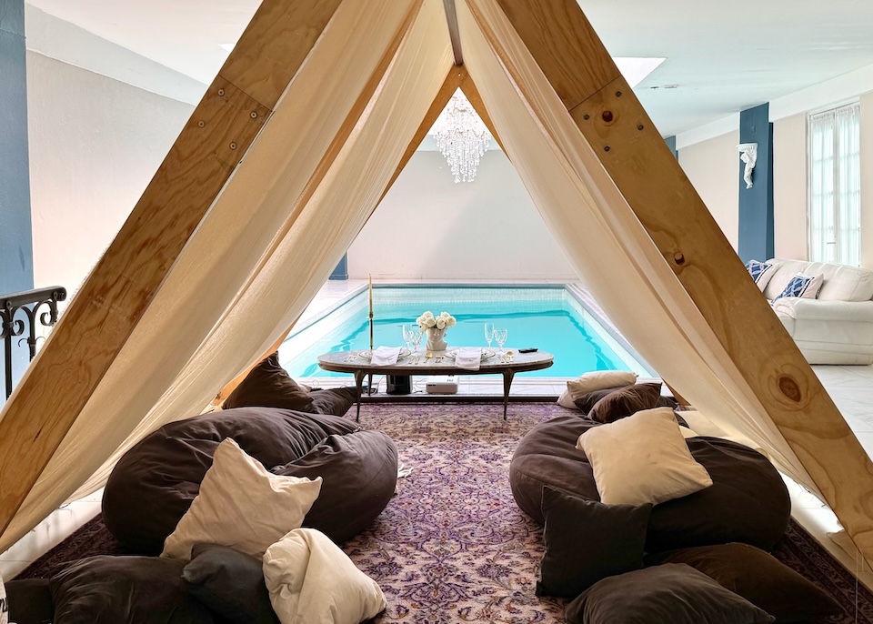 An A-frame poolside cabana set up with a Persian rug and pillows next to an indoor pool with a chandelier above at Mansion Papilio in Mexico City.