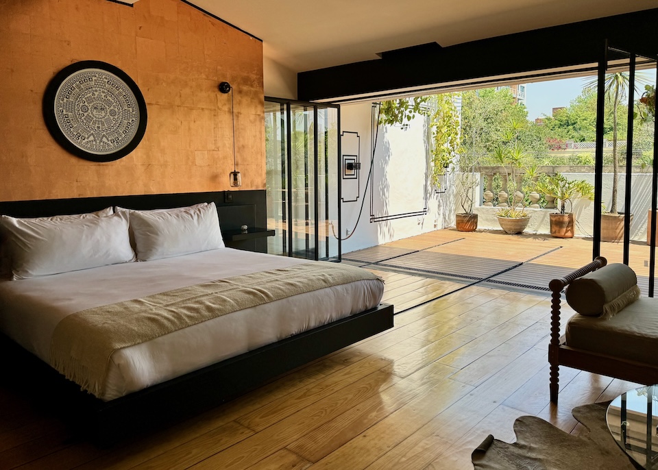 View from inside a suite with a king-sized bed mounted on tracks embedded into the floor that lead to a sunny terrace with plants at La Valise Hotel in Mexico City.