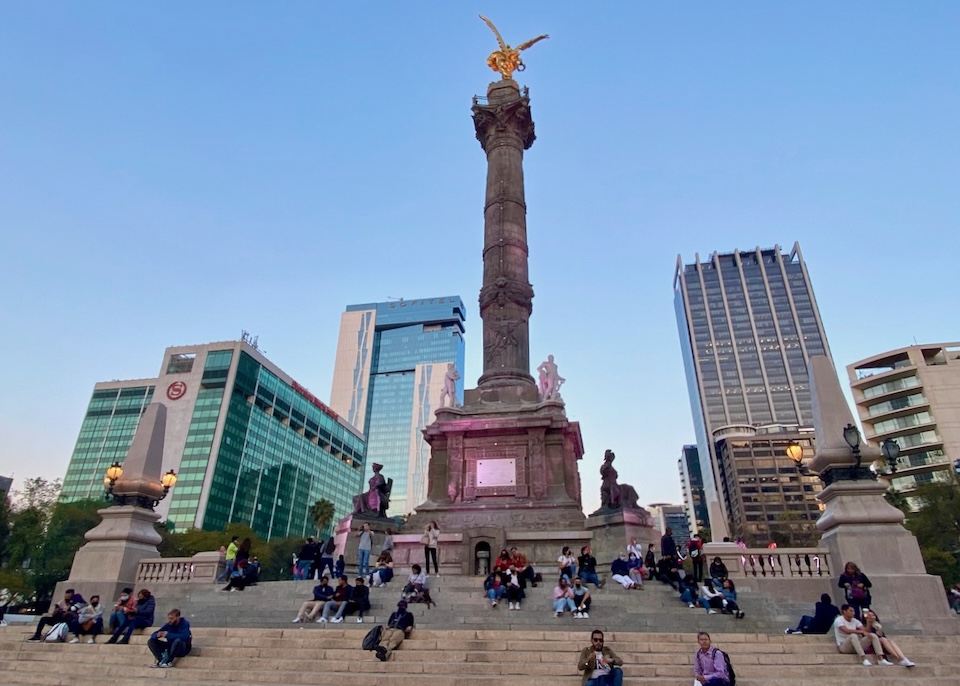 People sitting on the steps below the Angel of Independence monument at sunset with hotels in the background in colonia Cuauhtémoc in Mexico City.